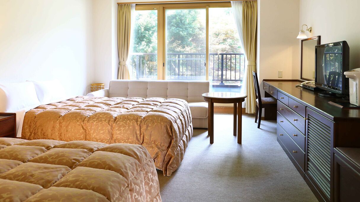 A hotel room with a large bed, a small table, a TV, and a balcony with a view of trees.