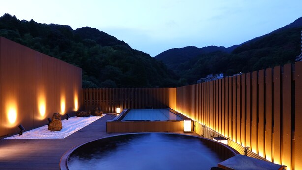A serene outdoor hot tub area with wooden decking, a large circular hot tub, and a wooden wall with integrated lighting. - Yugawara Chiyodasou (Yugawara)