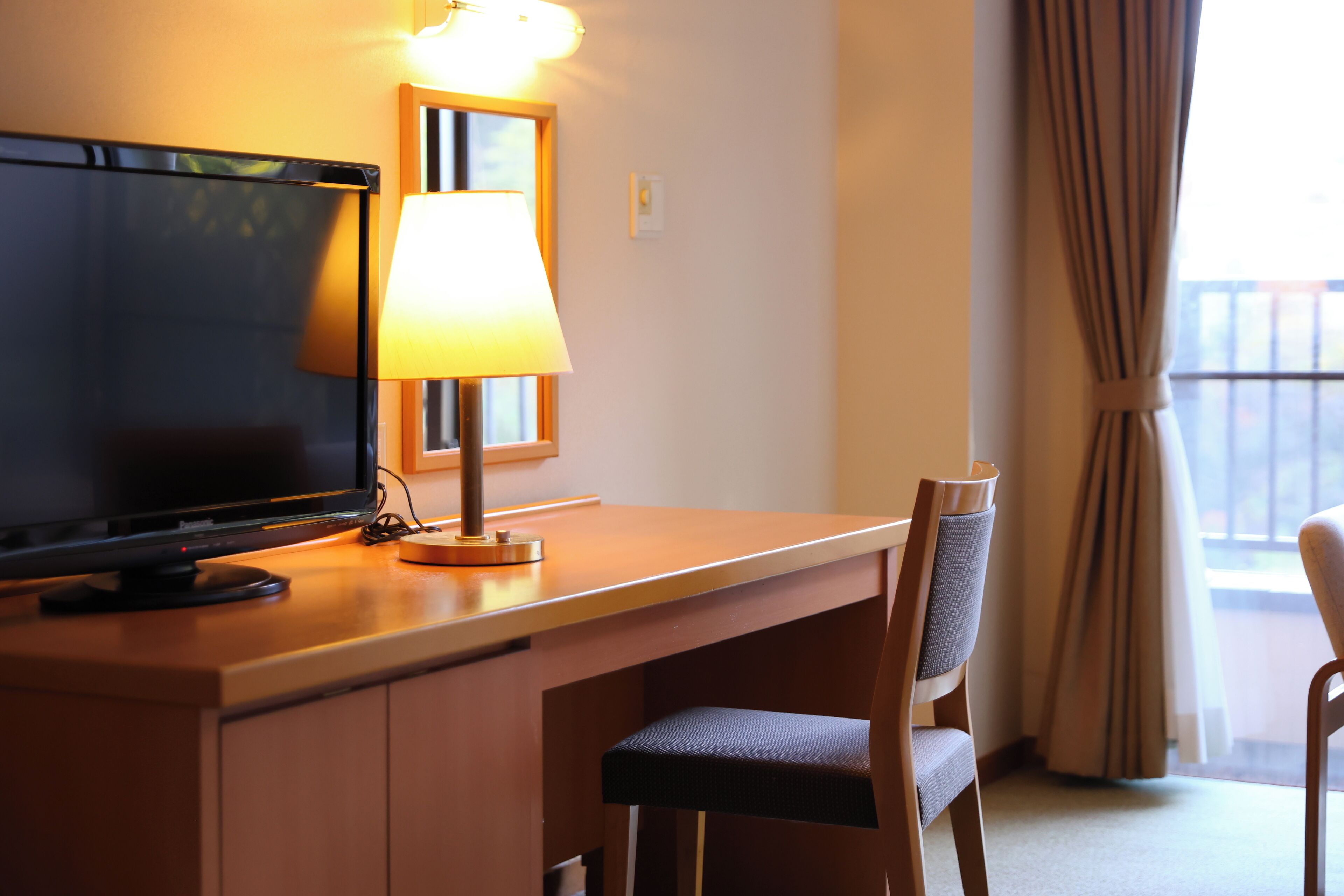 A hotel room with a desk, a television, a lamp, and a chair.