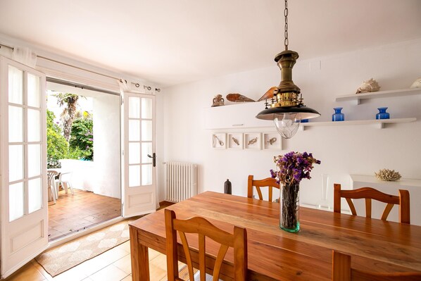 Un comedor con mesa de madera, sillas, un jarrón con flores, una lámpara colgante y vistas al jardín a través de puertas abiertas.