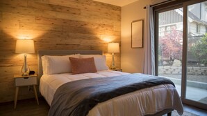 A bedroom with a wooden wall, a large bed with white bedding, two bedside lamps, and a view of a garden through a sliding glass door.
