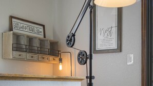 A cozy room with a wooden shelf displaying a sign, a black floor lamp, a framed artwork, and a textured ottoman.