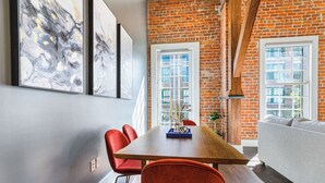 A dining area with a wooden table, red chairs, and a brick wall. There are two windows with white frames and a white sofa in the background. - 💙 DWNTWN NOLA CHARMER,WITH ALL THE FEELS! 2BR+QUEEN Sleeper Sofa+Private Balcony💙The Steamboat (New Orleans)