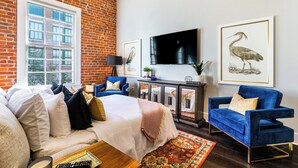 A living room with a brick wall, a large window, a sofa, a blue armchair, a TV, and framed artwork. - 💙 DWNTWN NOLA CHARMER,WITH ALL THE FEELS! 2BR+QUEEN Sleeper Sofa+Private Balcony💙The Steamboat (New Orleans)