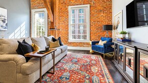 A living room with a brick wall, a sofa, a blue chair, a TV, and a patterned rug. - 💙 DWNTWN NOLA CHARMER,WITH ALL THE FEELS! 2BR+QUEEN Sleeper Sofa+Private Balcony💙The Steamboat (New Orleans)