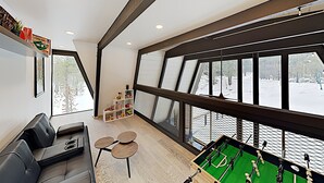 Une chambre moderne avec un canapé, une petite table et un baby-foot. De grandes fenêtres offrent une vue sur un paysage enneigé.