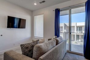 A modern living room with a flat-screen TV, a sofa with patterned cushions, and a large sliding glass door leading to an outdoor balcony.