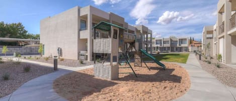 A playground with a slide and climbing structure, surrounded by a landscaped area with a concrete path.