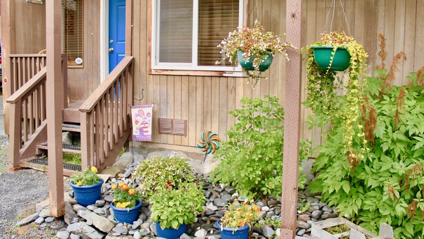 Una casa de madera con puerta azul, un porche con baranda de madera y varias plantas en macetas.