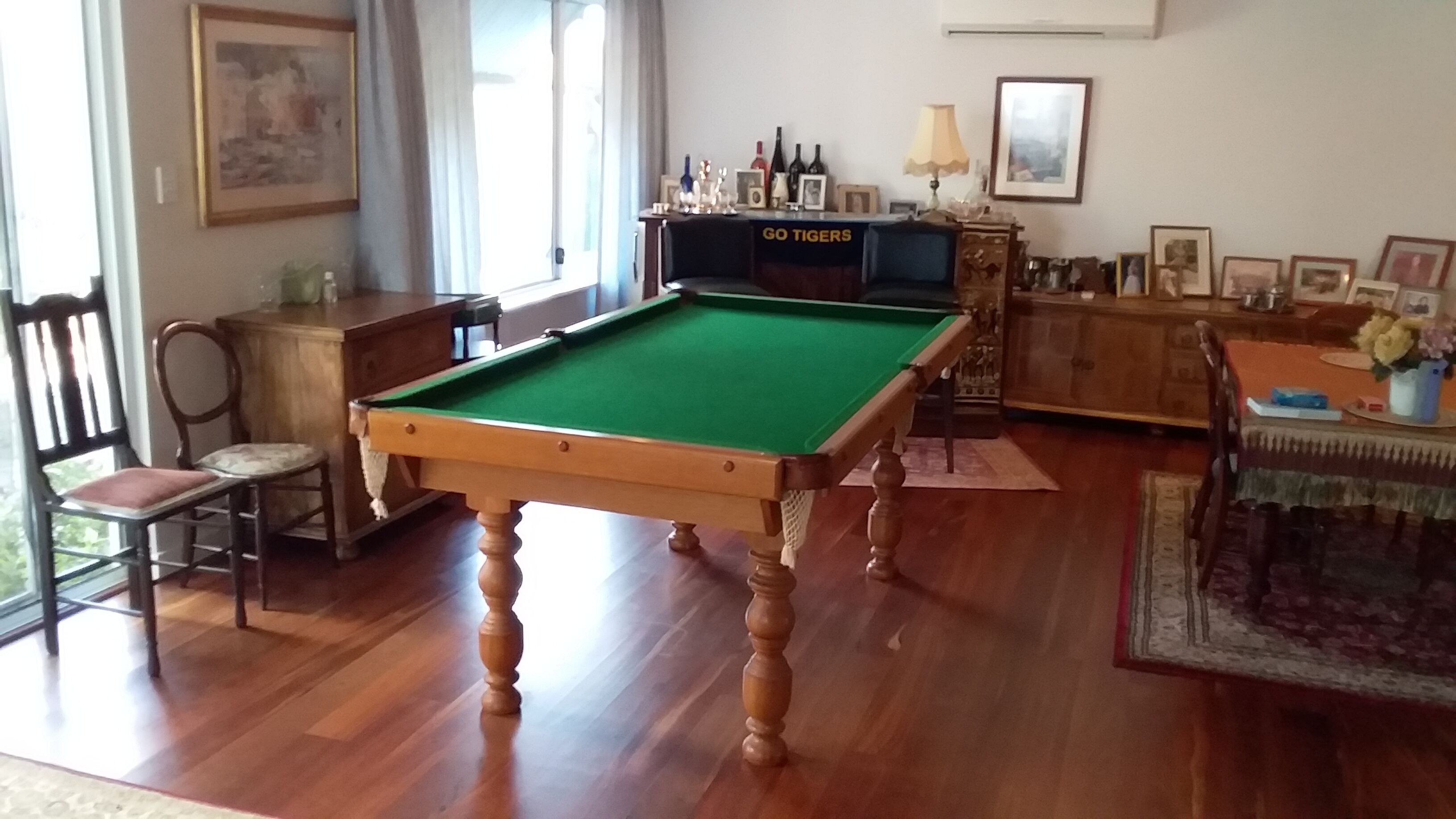 Una mesa de billar en una habitación con piso de madera, una silla, un armario y una mesa con lámpara.