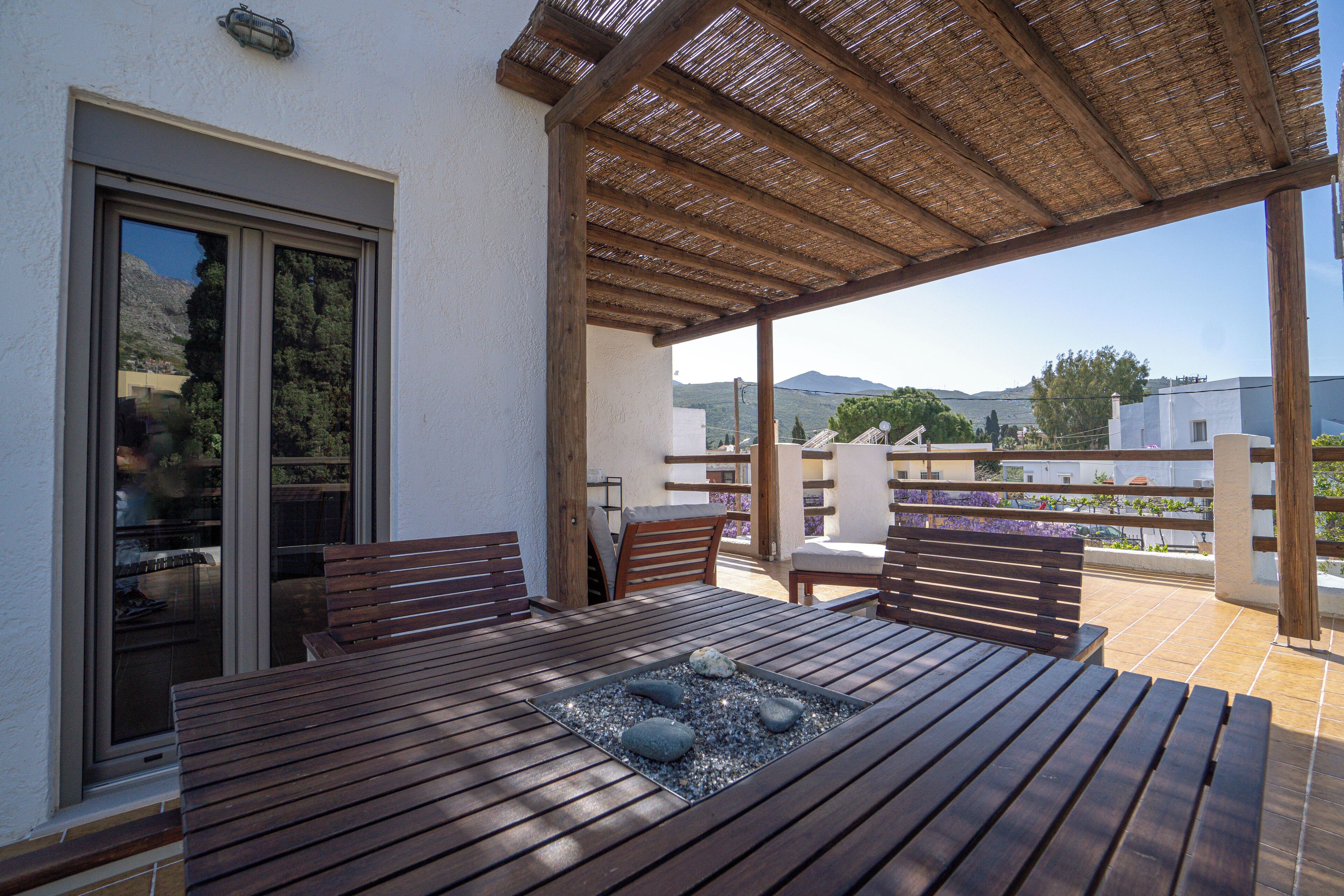 Une table en bois, accompagnée de quatre chaises et d’un plateau décoratif en verre, est placée sur un balcon offrant une vue sur les montagnes.