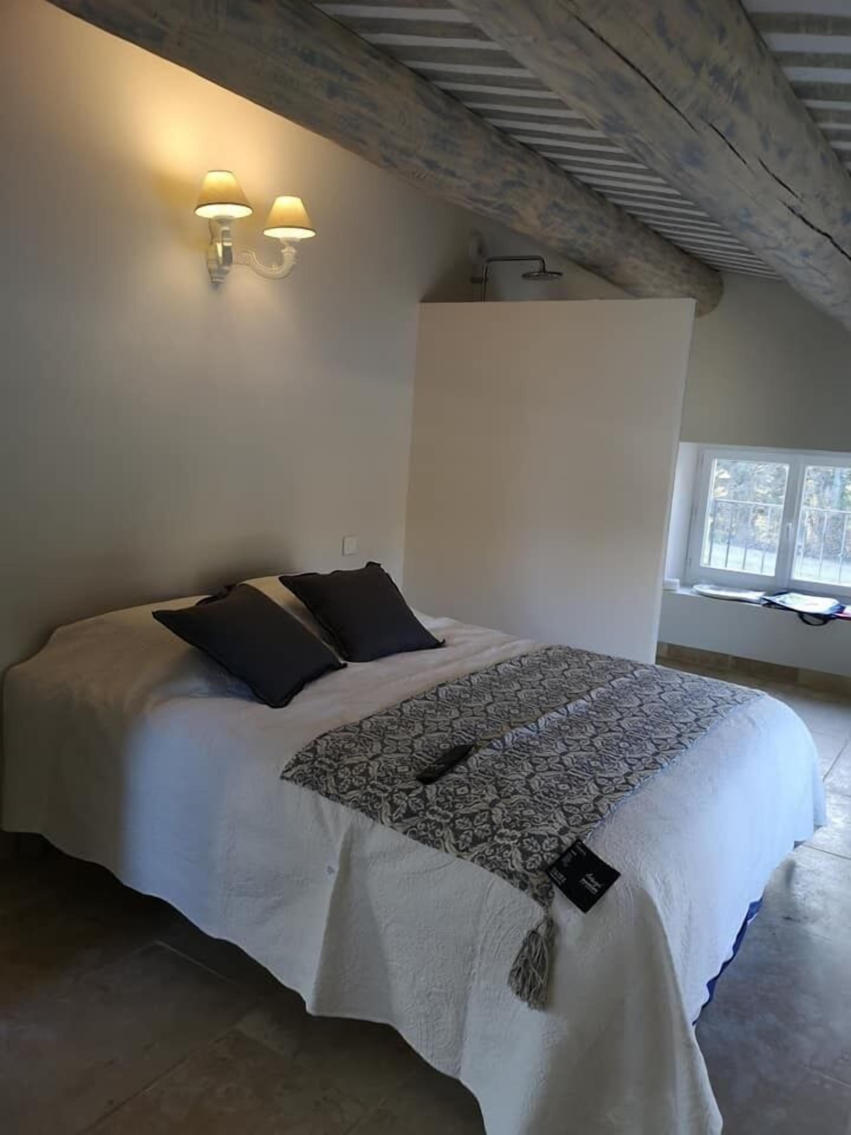 Une chambre à coucher avec un lit, deux lampes fixées au mur, une fenêtre et un plafond avec des poutres apparentes.