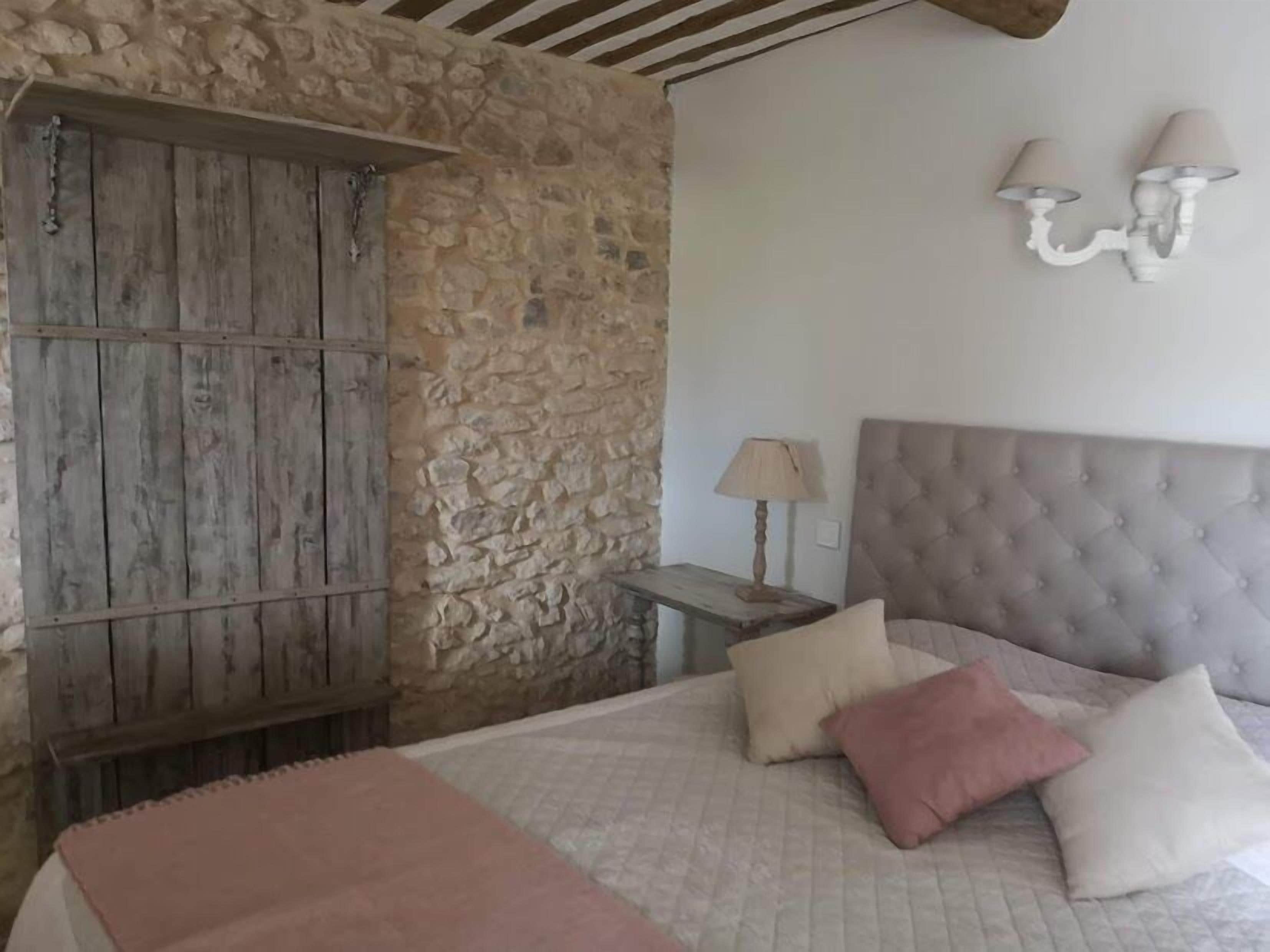 Un dormitorio con pared de piedra, puerta de madera, cama con cabecera capitonada y dos lámparas fijadas a la pared.
