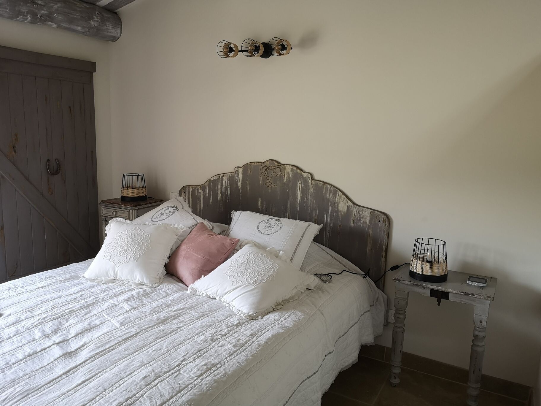 A bedroom with a wooden headboard, a bed with white bedding and decorative pillows, a bedside table with a lamp, and a rustic wall decor.