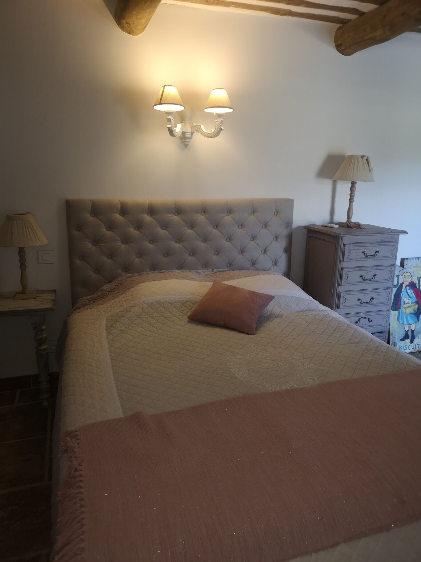 A bedroom with a tufted headboard, a bed with a quilt and a single pillow, a nightstand with a lamp, and a dresser with a lamp.