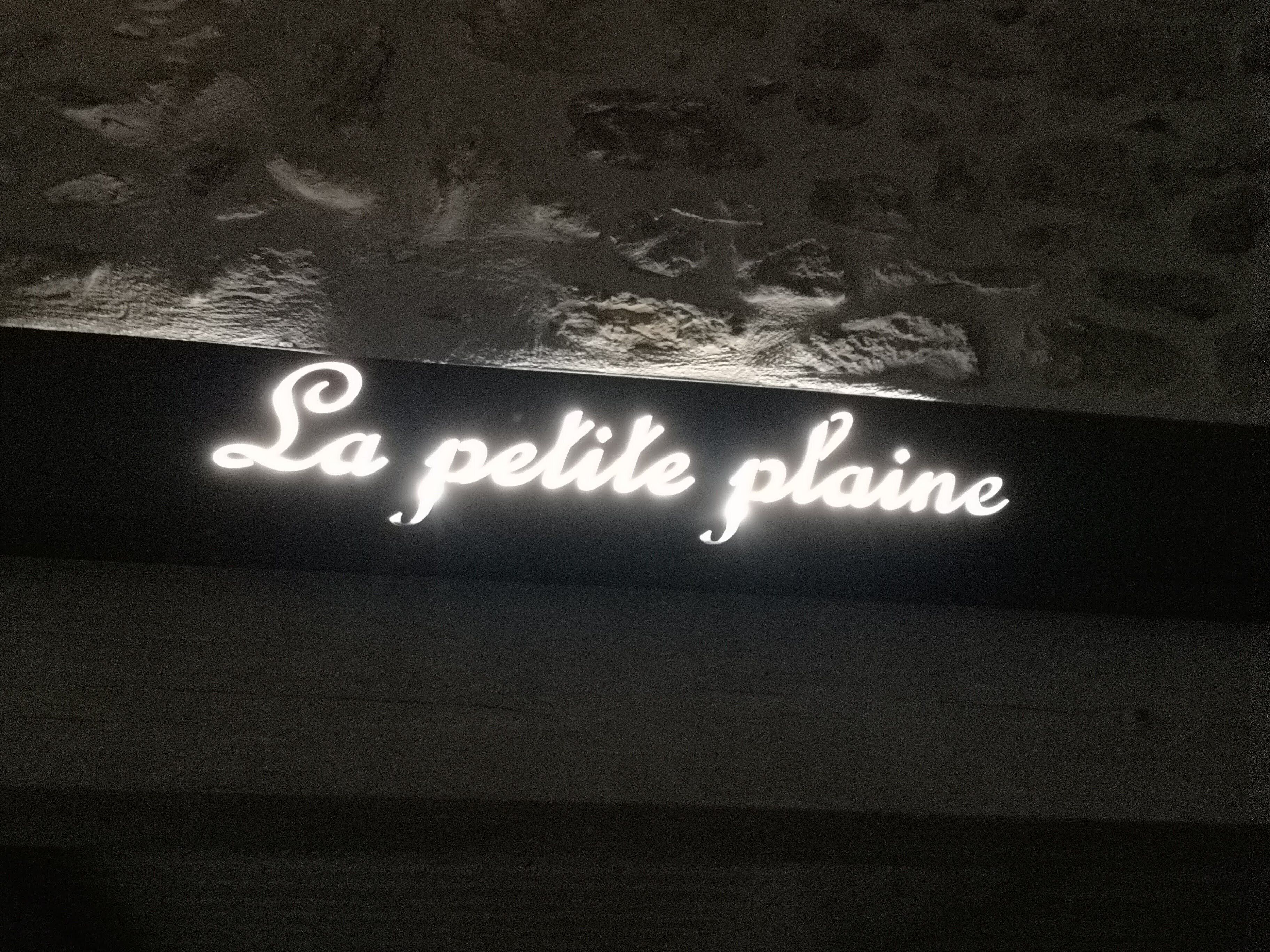Neonvalomainos, jossa lukee ”La petite plaine”, ja taustalla kiviseinä.