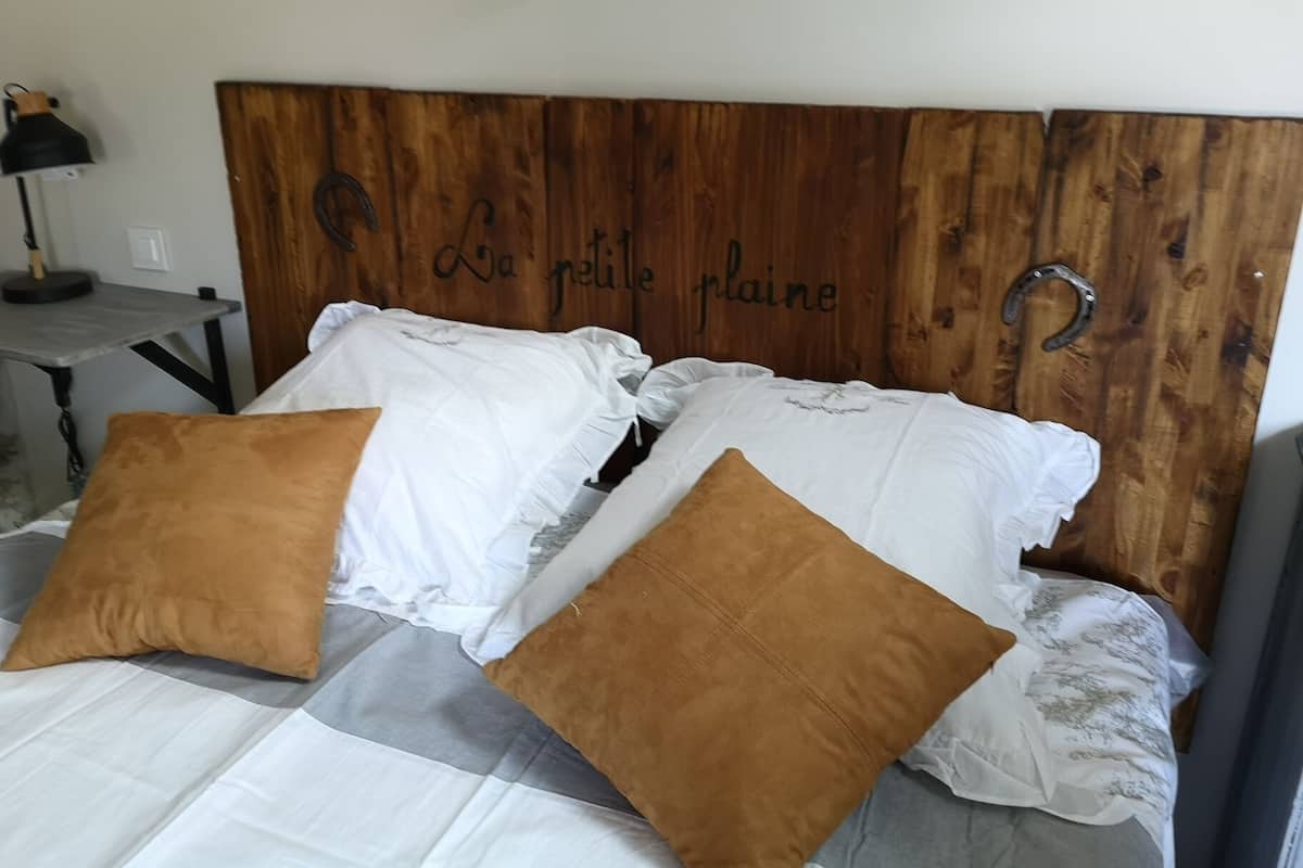 Una cama con almohadas blancas y marrones, un cabecero de madera con el texto "la petite prairie", y una mesita de noche con una lámpara a la izquierda.