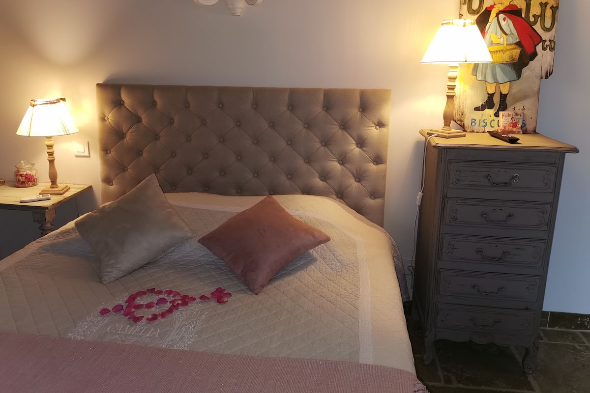Un dormitorio con cabecera capitonada, una mesita de noche con lámpara, un aparador con cajones y una pared con un cuadro.