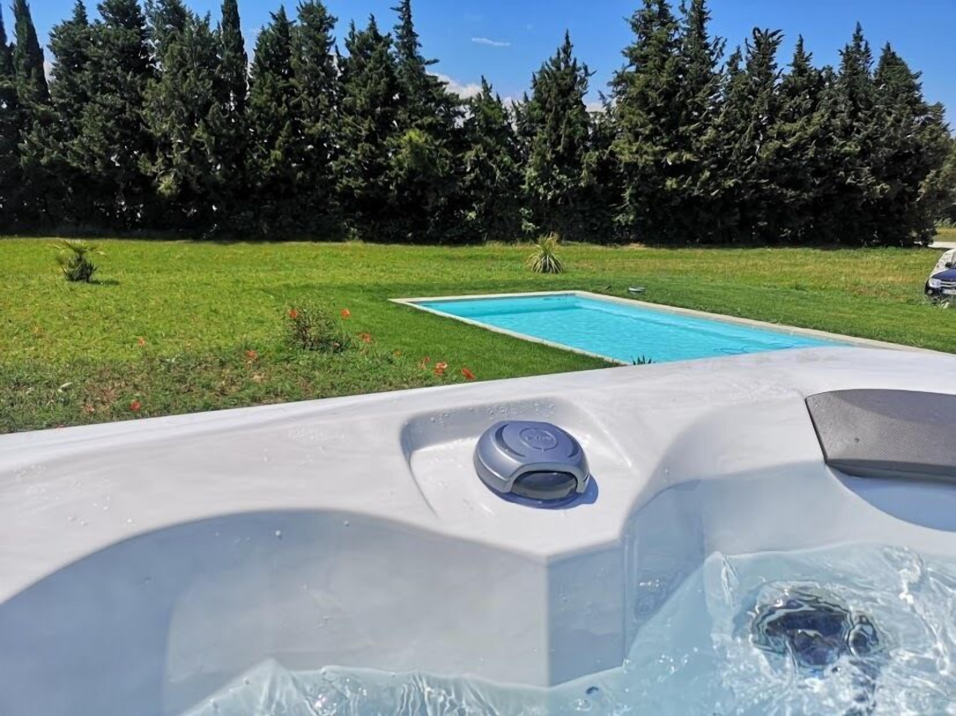 A hot tub with a control panel, surrounded by a grassy area and a swimming pool, with trees in the background.
