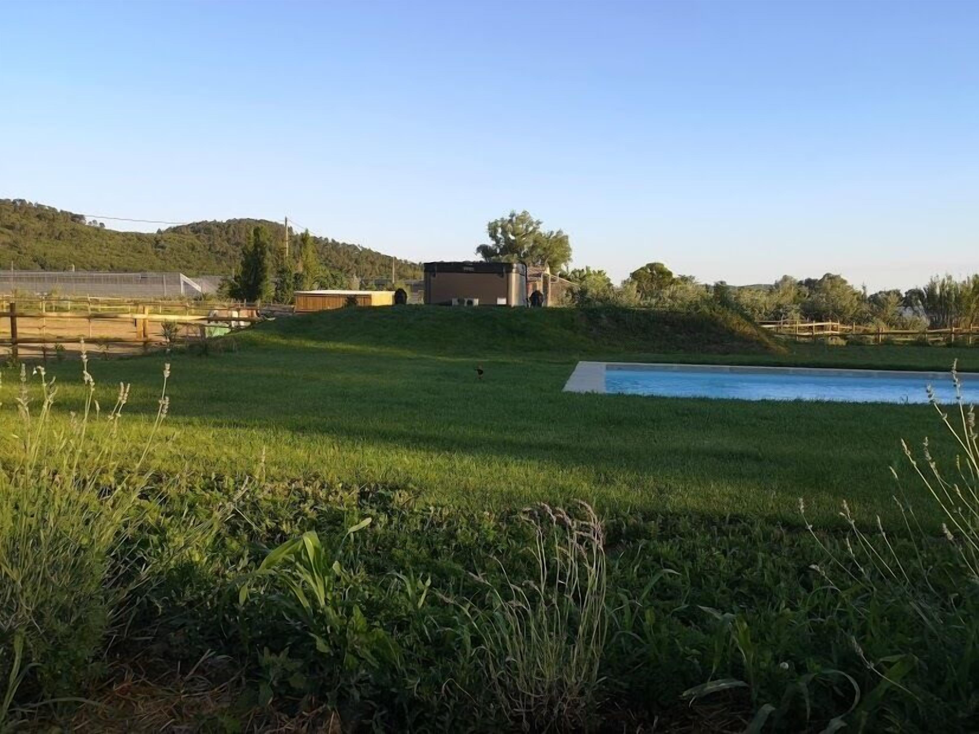 Uma área gramada com piscina, um cercado e edifícios ao fundo.