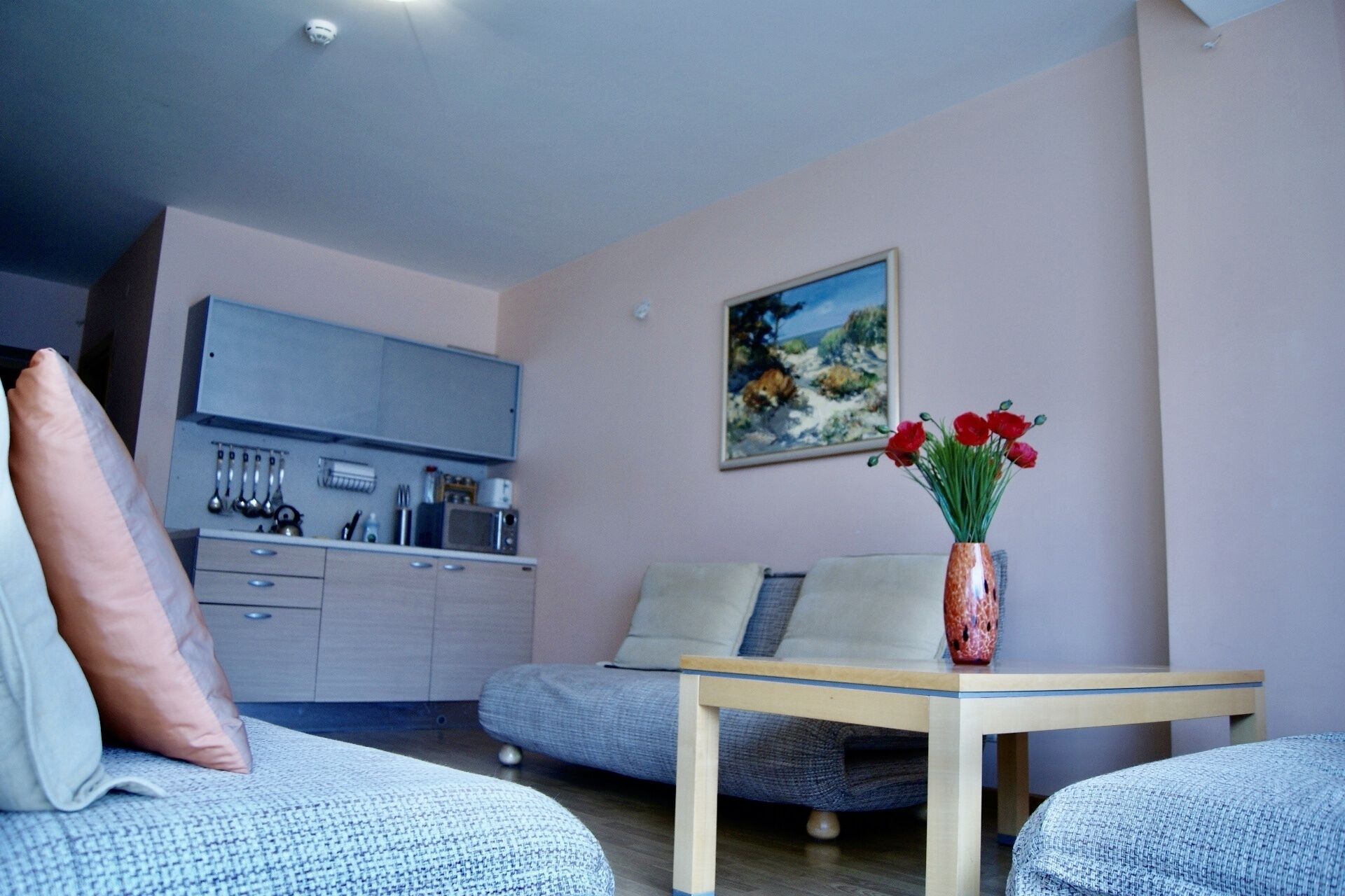 Un salón moderno con sofá, una mesita con un jarrón de flores, una zona de cocina con armarios y microondas, y un cuadro en la pared.