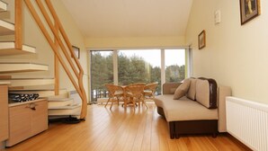 Amplia sala de estar con una escalera de madera, un sofá, una mesa de comedor con sillas y grandes ventanas con vistas a árboles.