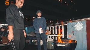 Two people standing near a large outdoor grill with flames, in an urban setting with buildings in the background.