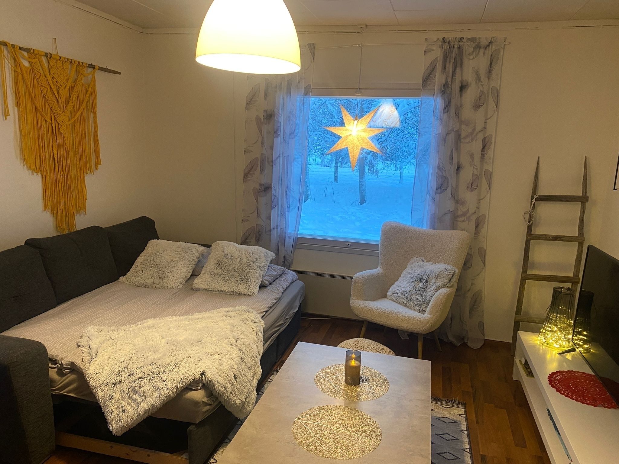 A cozy living room with a sofa, armchair, and a table with a candle. A star decoration is visible through the window.