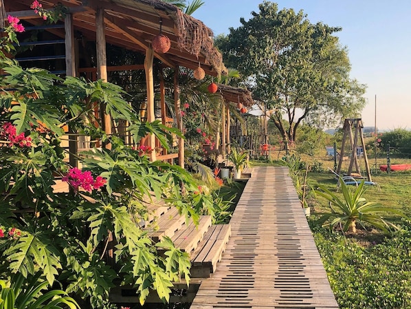 A wooden deck leading to a covered area with a thatched roof, surrounded by lush greenery and flowering plants.