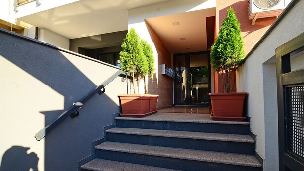 Una serie de escalones exteriores que conducen a la entrada de un edificio, flanqueados por dos árboles en macetas y un equipo de aire acondicionado grande.