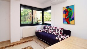Una sala de estar moderna con sofá, mesa de comedor, radiador, ventana con vista a follaje y un cuadro abstracto colorido en la pared.