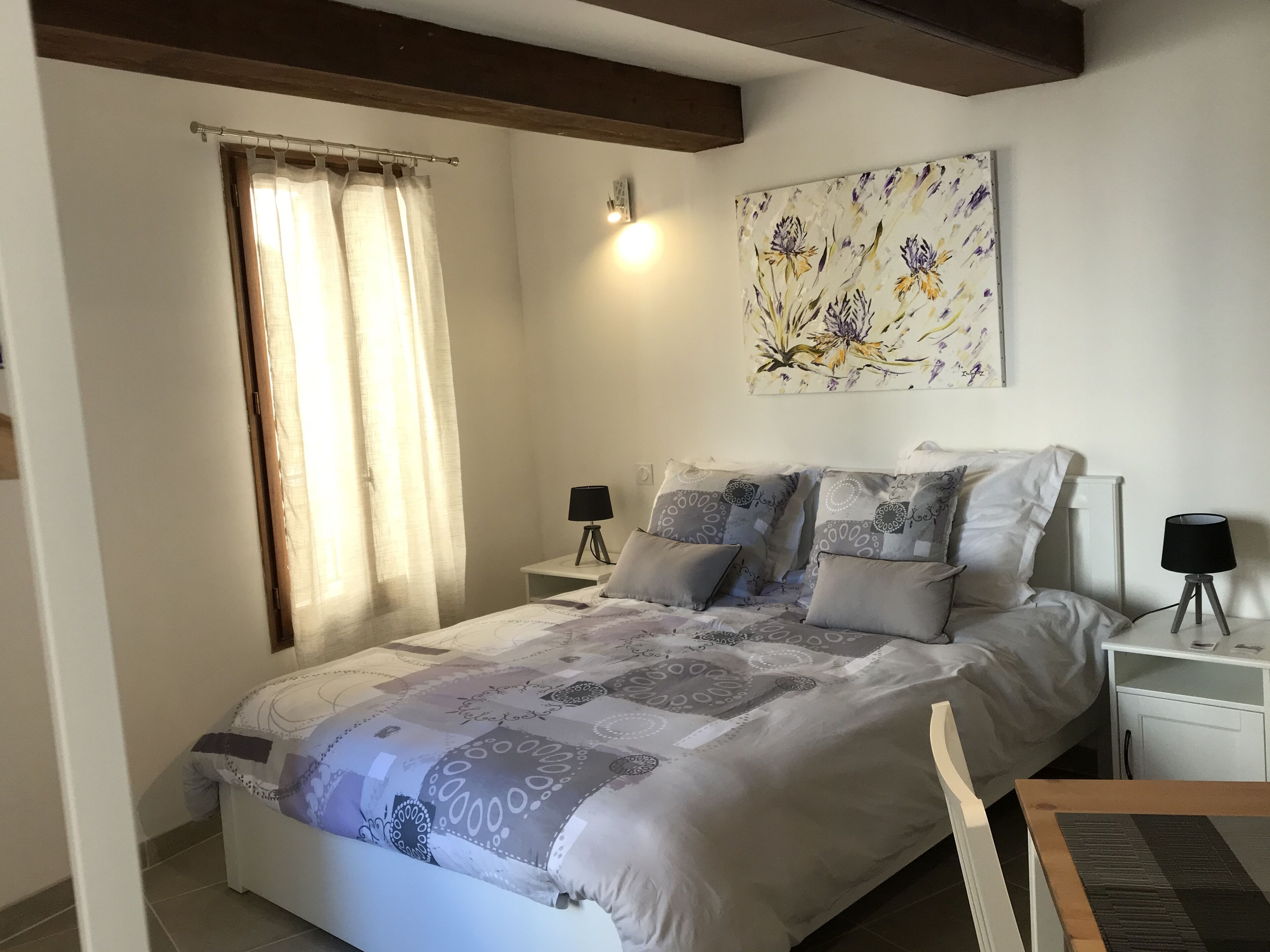 Une chambre à coucher avec un lit, deux tables de chevet, une lampe sur chaque table, une fenêtre avec des rideaux et un tableau floral accroché au mur.