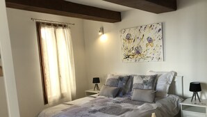 A bedroom with a bed, two bedside tables, a lamp on each table, a window with curtains, and a floral painting on the wall.