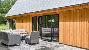 Ein modernes Gebäude mit Holzfassade und großen Glasschiebetüren. Im Inneren befindet sich eine Sitzecke mit Korbmöbeln und einem kleinen Tisch.