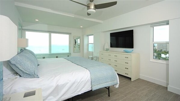 A bedroom with a bed, dresser, TV, and large windows.