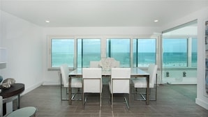 Un comedor con mesa y sillas de vidrio, ventanas amplias con vista al océano y un adorno blanco sobre la mesa.