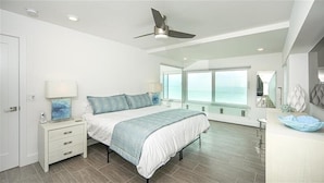 Un dormitorio moderno con cama, mesitas de noche, ventilador de techo y ventanas amplias con vistas al mar.
