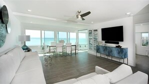 Una sala de estar moderna con un sofá blanco, un comedor con mesa y sillas, un televisor grande montado en un mueble azul y un ventilador de techo.