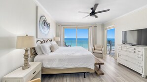 Habitación de hotel con cama, televisión, ventilador de techo, balcón con vista al océano y cómoda.