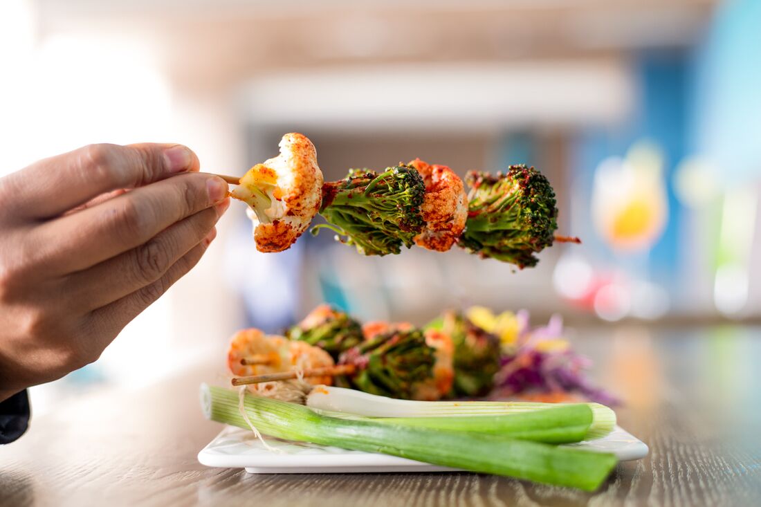 A hand holding a piece of food, possibly a vegetable skewer, with broccoli and cauliflower visible.