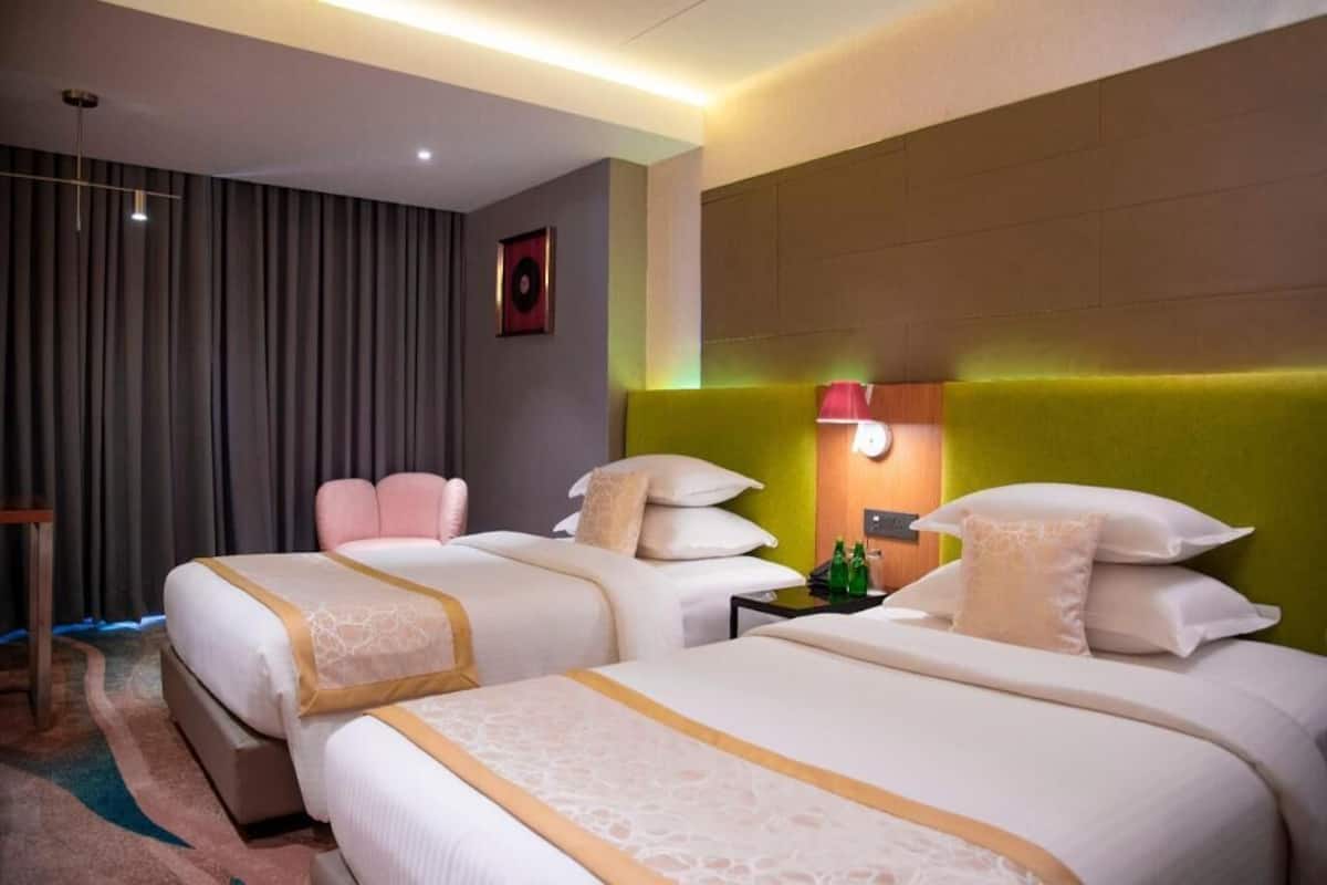 Habitación de hotel con dos camas, cabecera, mesita de noche, lámpara y cortina.