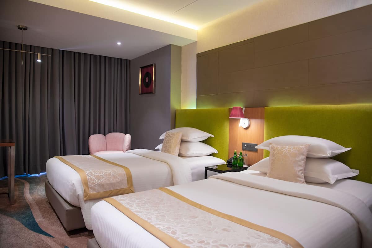 Habitación de hotel con dos camas, un cabecero, una mesita de noche con lámpara y un reloj de pared.