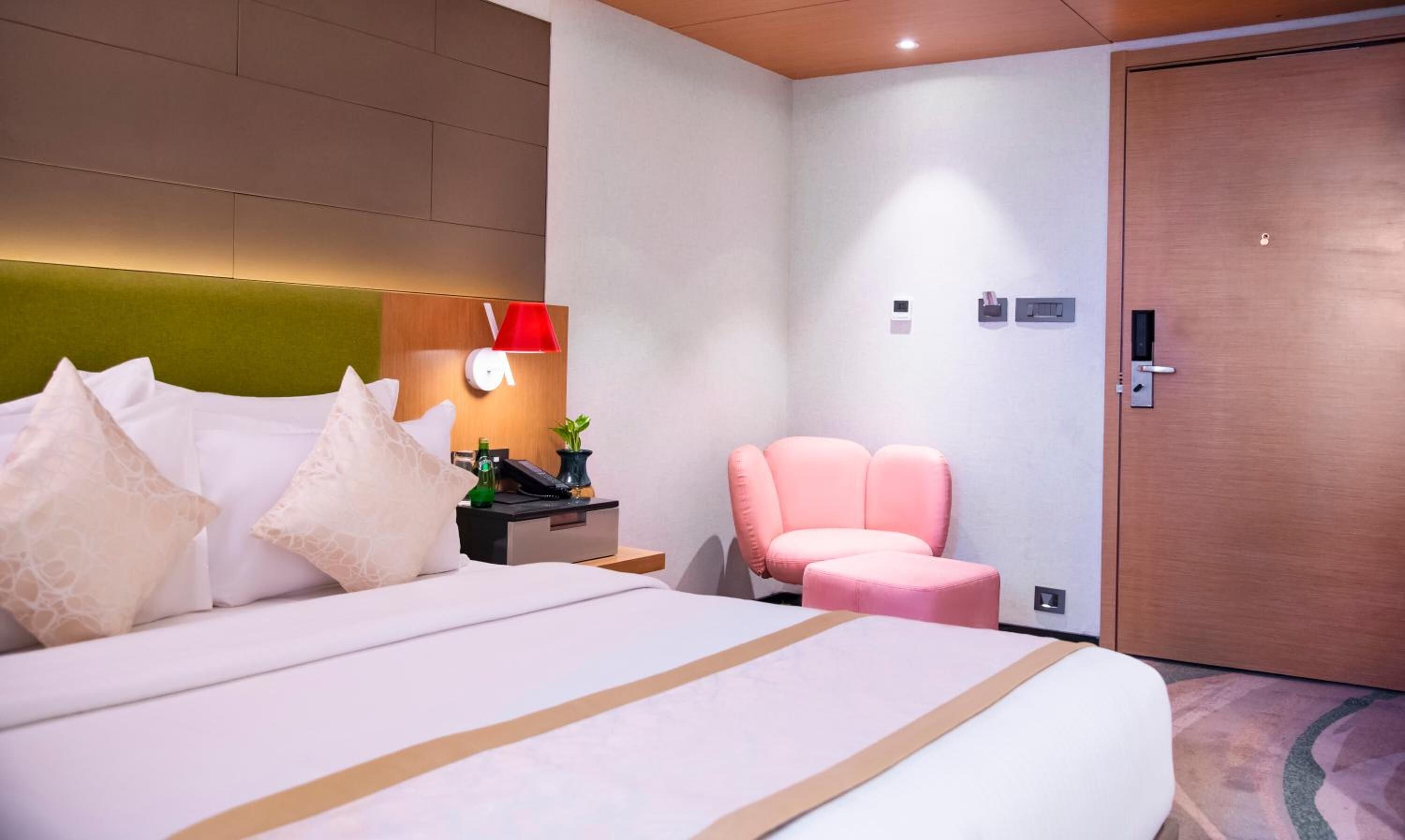 Una habitación de hotel con una cama grande, una mesita de noche con lámpara, una silla rosa y una puerta con cerradura.
