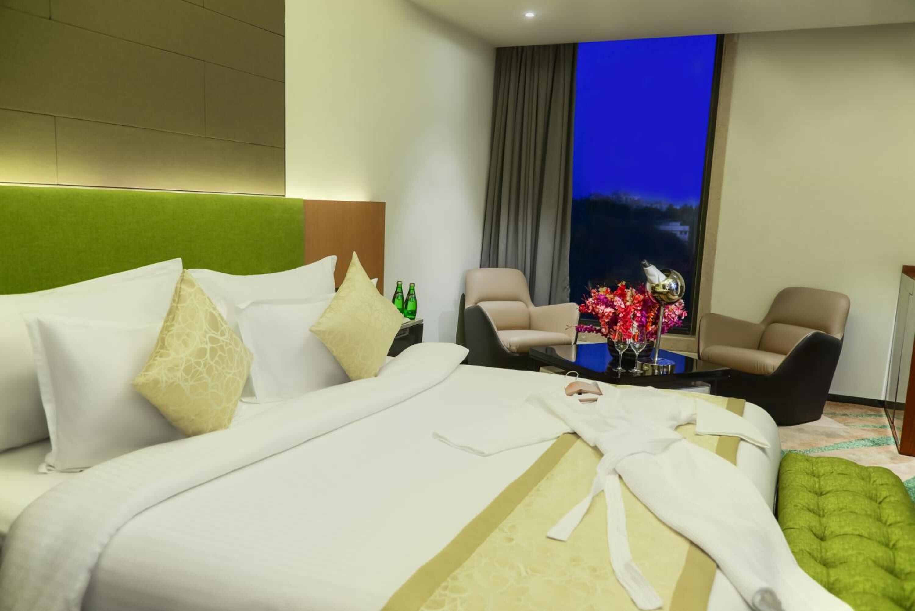 Habitación de hotel con una cama grande, dos sillones, una mesita con un florero y vistas al cielo nocturno.