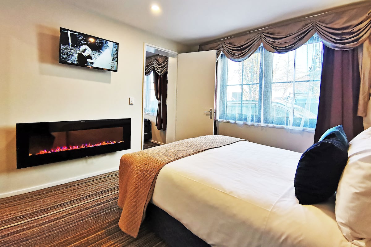 A hotel room with a bed, a flat-screen TV mounted on the wall, a fireplace, and a window with curtains.
