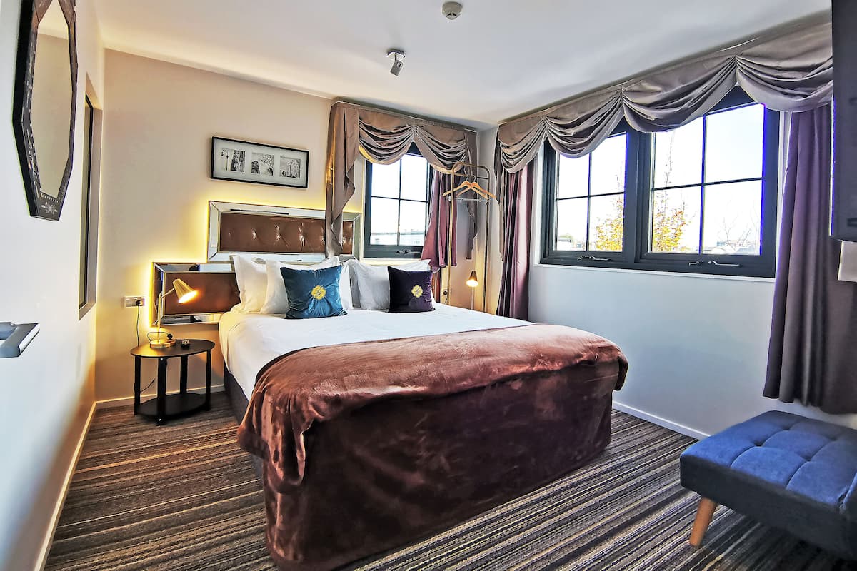 A hotel room with a large bed, a bedside table with a lamp, a window with curtains, and a small stool.