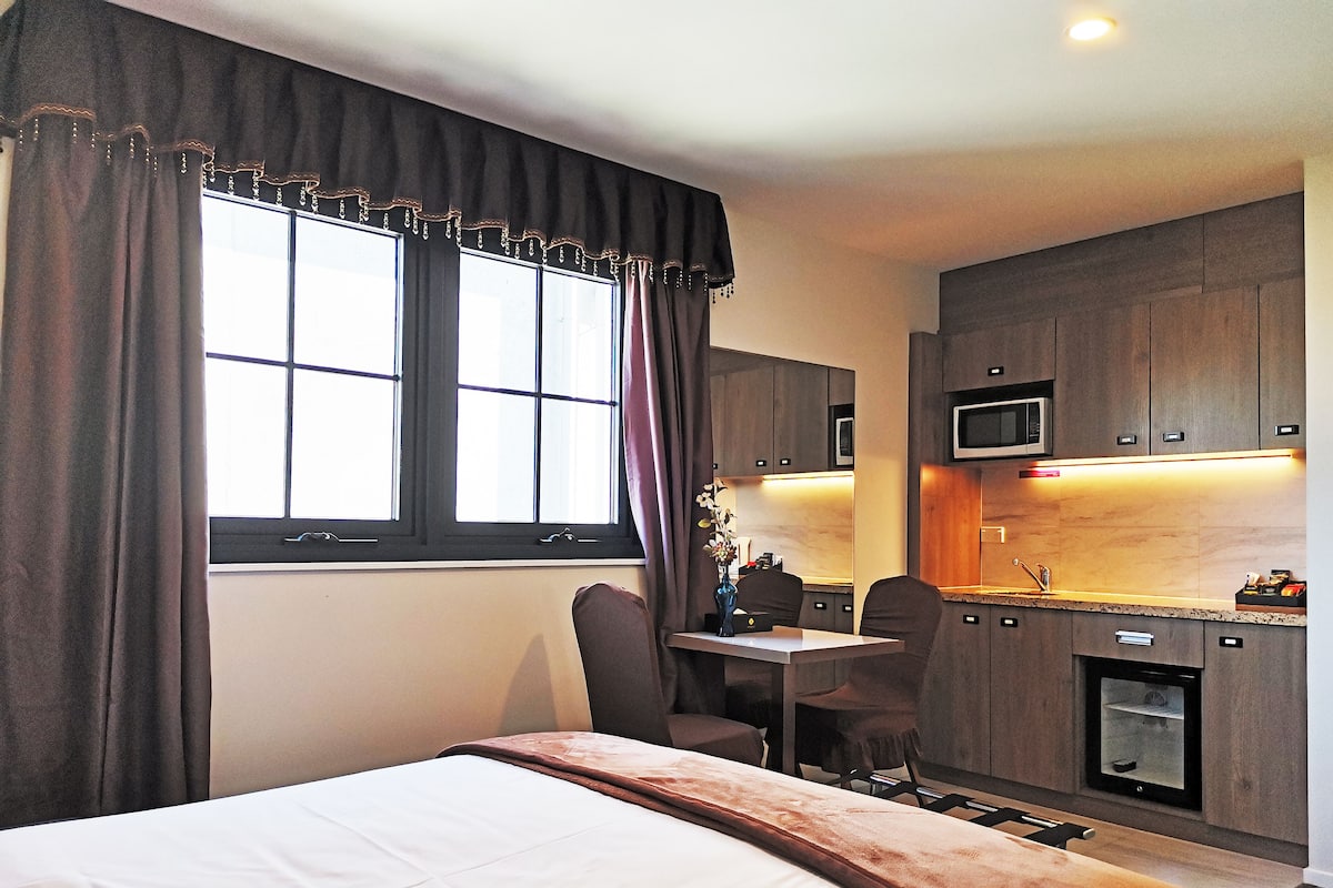 A hotel room with a bed, a desk, two chairs, a small kitchenette, and a window with curtains.