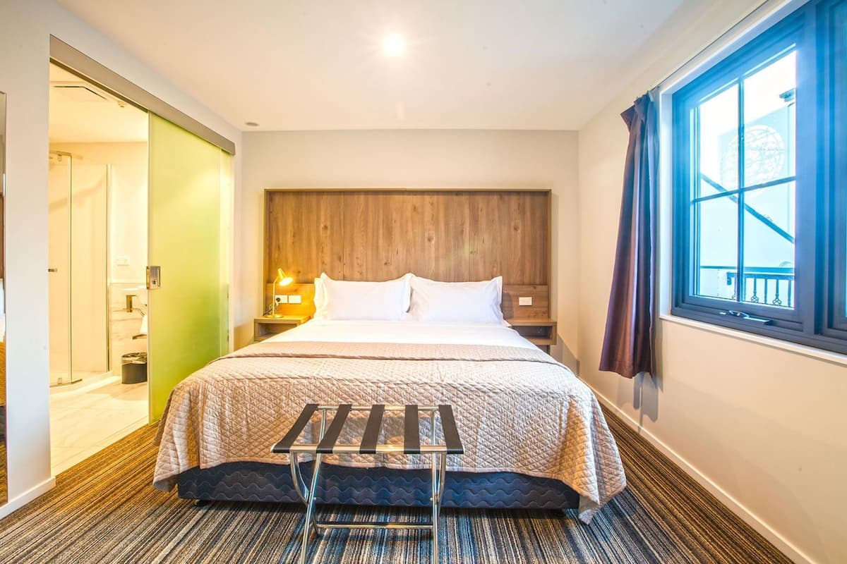 A hotel room with a bed, bedside tables, a nightstand, a window with curtains, and a door leading to a bathroom.