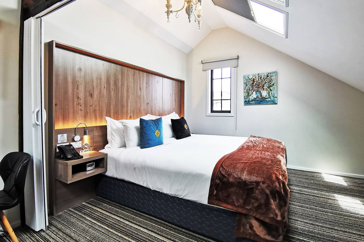 A hotel room with a large bed, a wooden headboard, a desk with a telephone, a chair, a window, and a painting on the wall.