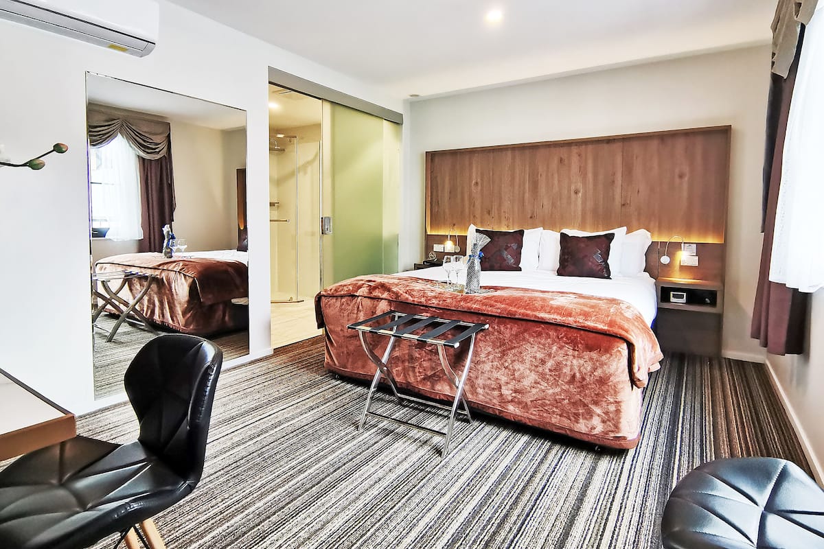 A hotel room with a large bed, a bedside table with a lamp, a mirror, a chair, and a small table with a glass of water.